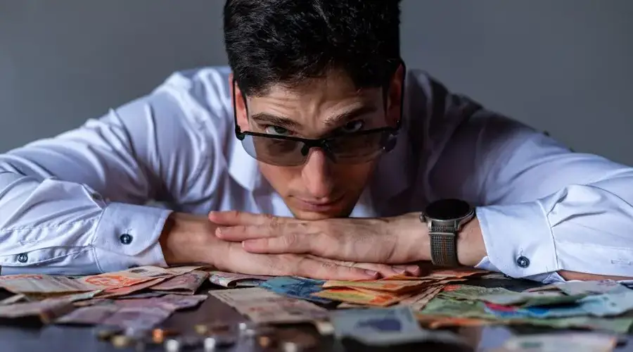 A man wearing glasses rests his head on his arms with Argentine peso notes spread on the table