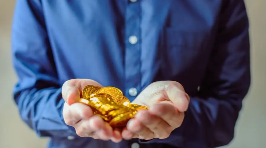 A person in a blue shirt holds a handful of shiny golden coins, symbolizing wealth and cryptocurrency.