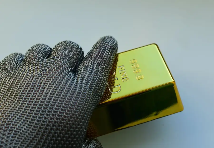 Close-up of a metal gloved hand holding a shiny gold bar indoors.