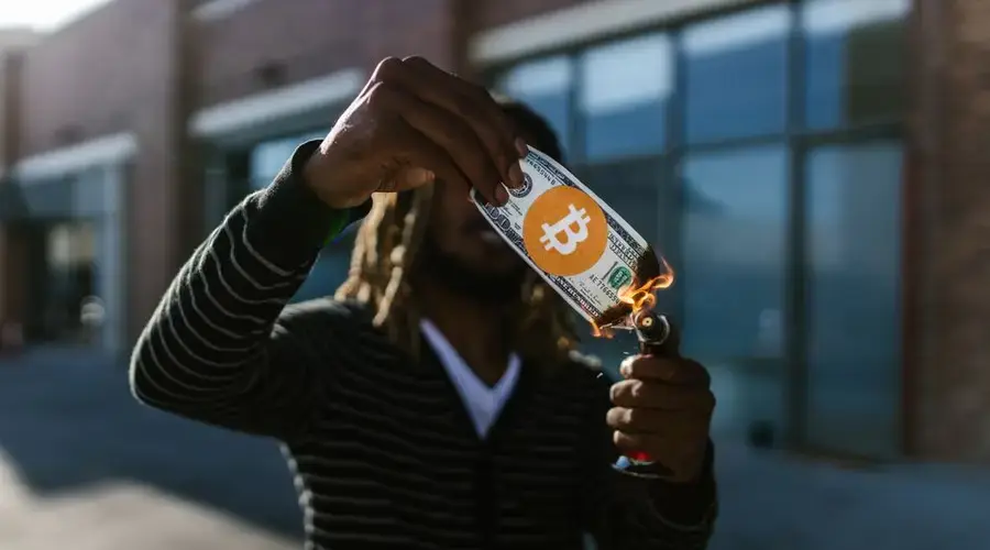 A man burns a fake dollar with a Bitcoin symbol outdoors, symbolizing cryptocurrency risks.