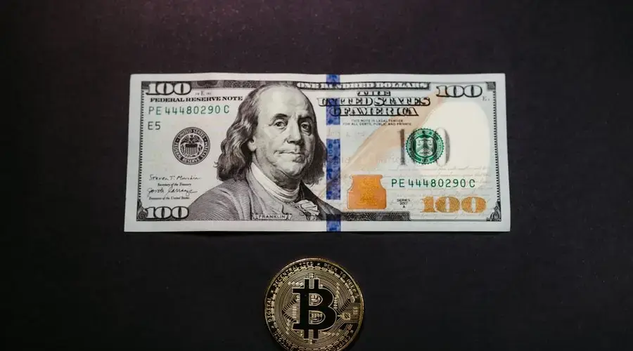 Image of a US 100 dollar bill and a Bitcoin coin on a black background, symbolizing traditional and digital currency.
