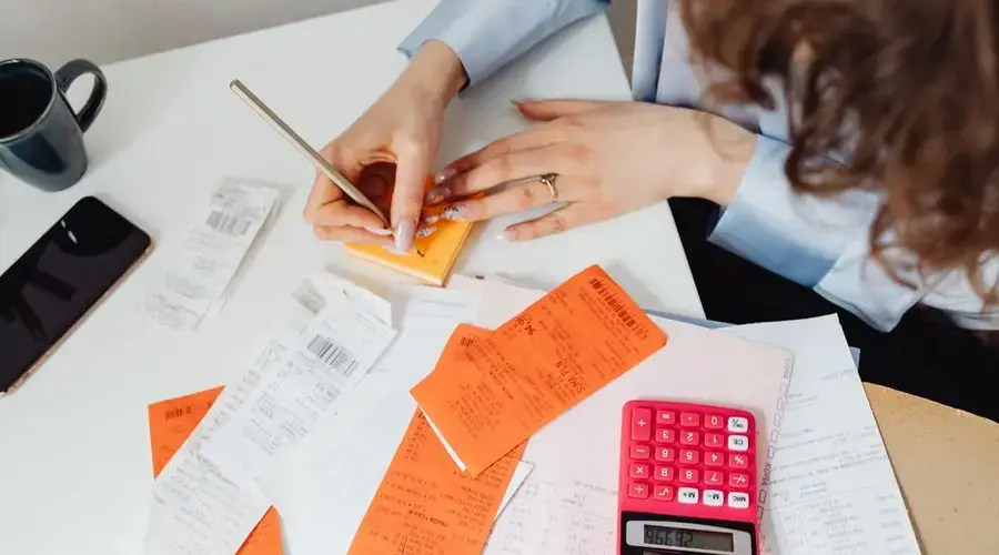 A woman writes down financial details using a calculator, receipts, and documents.