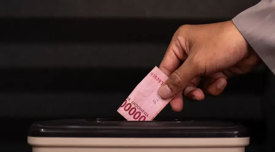 Close-up of a hand placing a 100,000 Indonesian Rupiah note into a donation box, highlighting charity and giving.