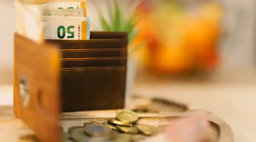 Brown wallet holding banknotes with scattered coins on a wooden table.