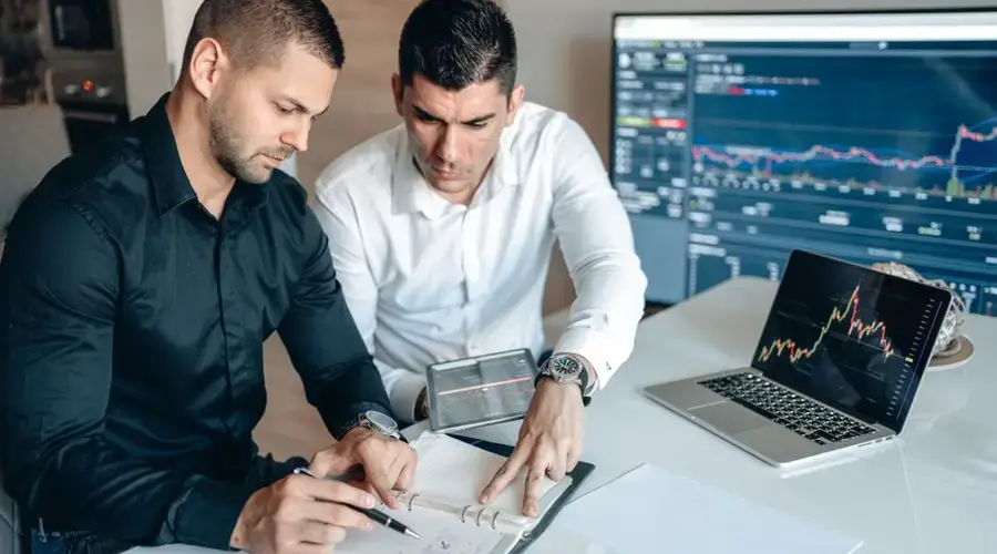 Two businessmen analyzing financial charts on digital devices in a modern office setting.