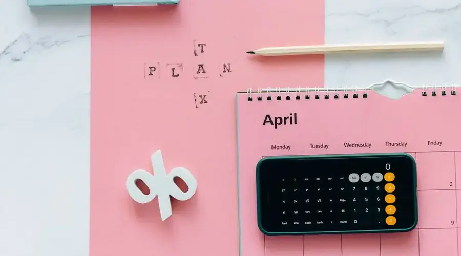 Flat lay of April calendar with calculator, pencil, and pink background for financial planning.