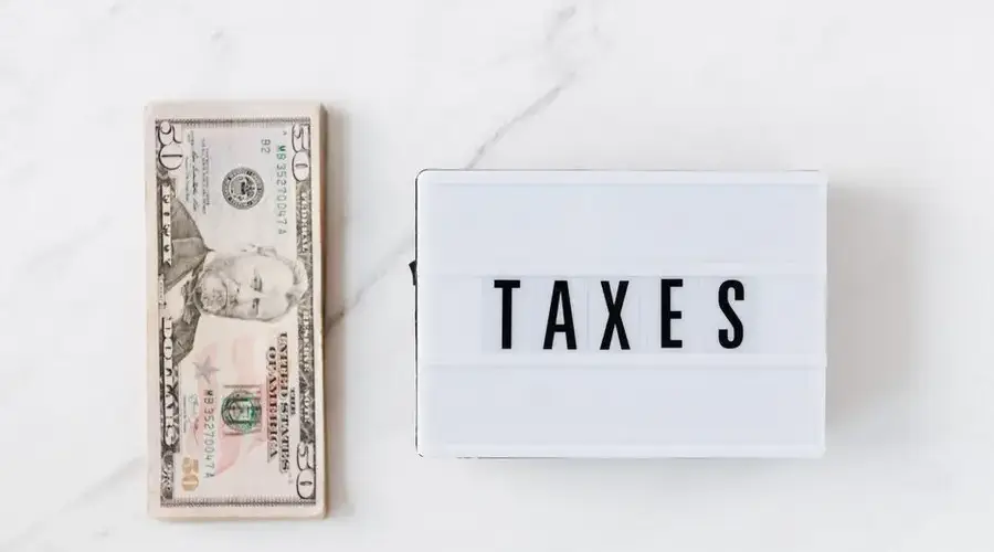 Top view composition of stack of American dollars placed on white marble surface with white retro light box with TAXES inscription