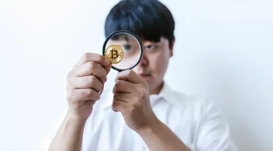 Close-up of a man examining a Bitcoin coin with a magnifying glass, symbolizing cryptocurrency analysis.
