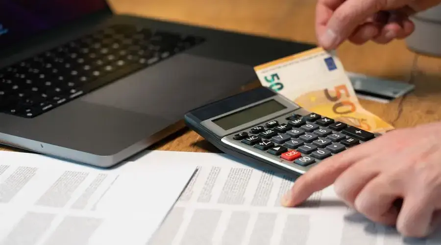Close-up of hands using calculator with euro notes and laptop on desk.