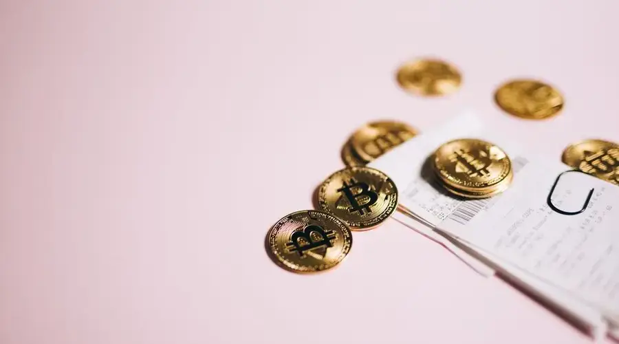 Close-up of Bitcoin coins and cash receipts on a soft pink background.