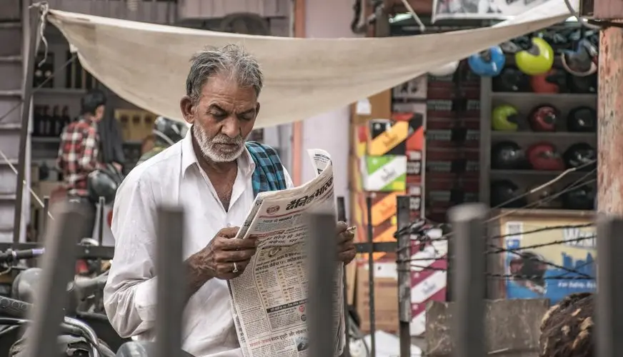 Elderly man immersed in a newspaper amidst a bustling urban street market.