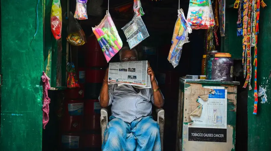 An elderly man reading a newspaper in a vibrant market shop filled with hanging goods.