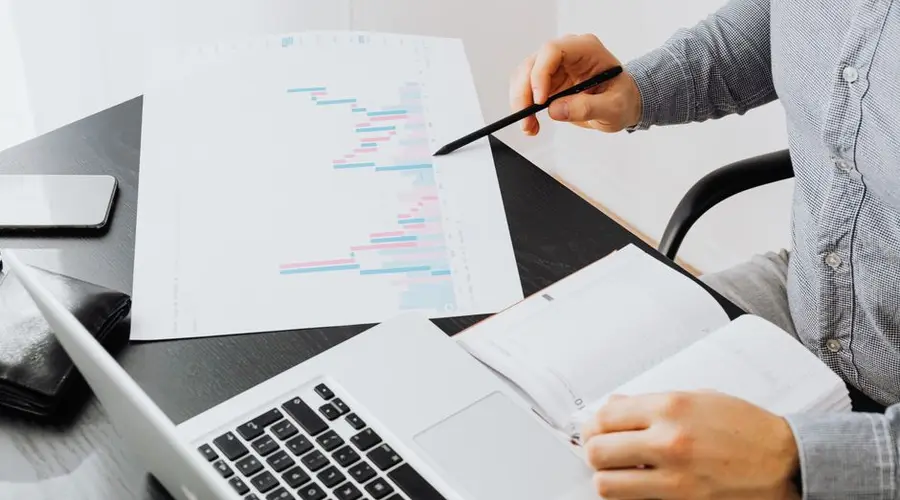 An office setup with a person analyzing a graph printout while using a laptop and notebook.