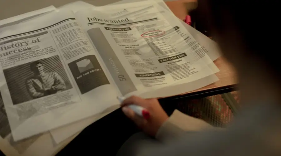 Person reviewing job listings in a newspaper, holding a pink marker for highlighting job ads.
