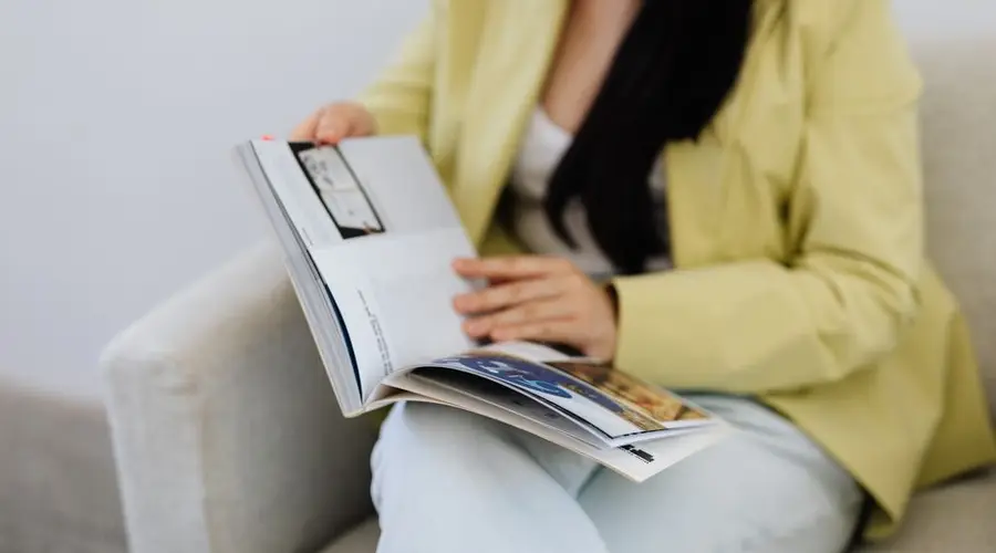 A woman in a yellow blazer reads a magazine on a comfortable chair indoors.