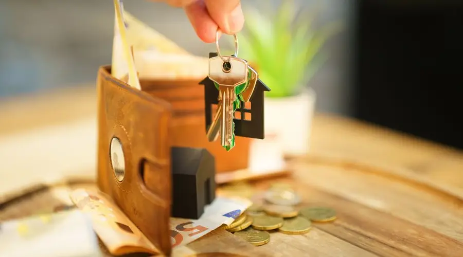 Close-up of hand holding a house key with a wallet and coins, symbolizing real estate investment.