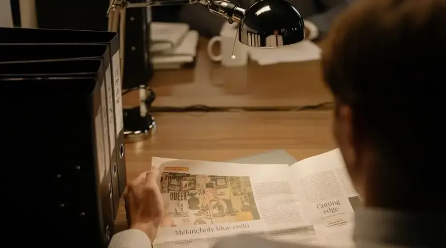 A person reads a newspaper at a desk with a lamp, suggesting investigation work.