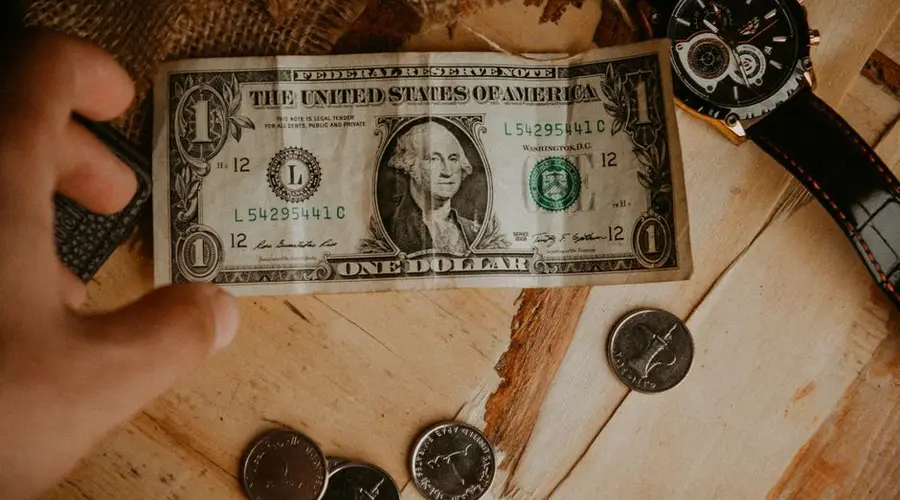 A person reaches for a one dollar bill surrounded by coins and a watch on a wooden table.