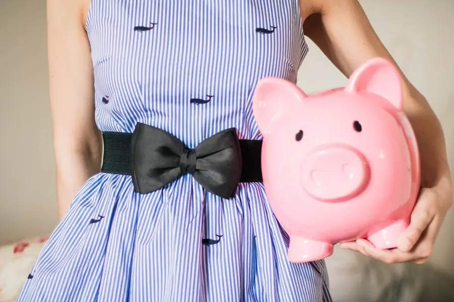 Crop unrecognizable female in lush striped dress with bow on belt with pink piggy bank in hand