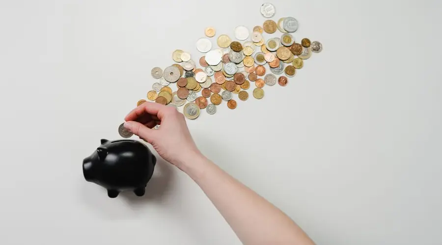 Person placing coin into a black piggy bank with scattered coins, symbolizing savings and finance.