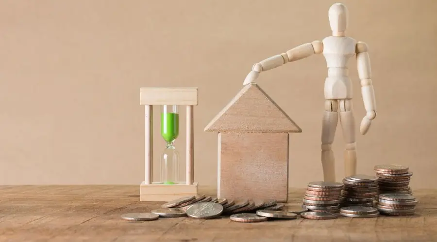 Wooden mannequin with house model, coins, and hourglass on a wooden table, symbolizing time and financial growth.