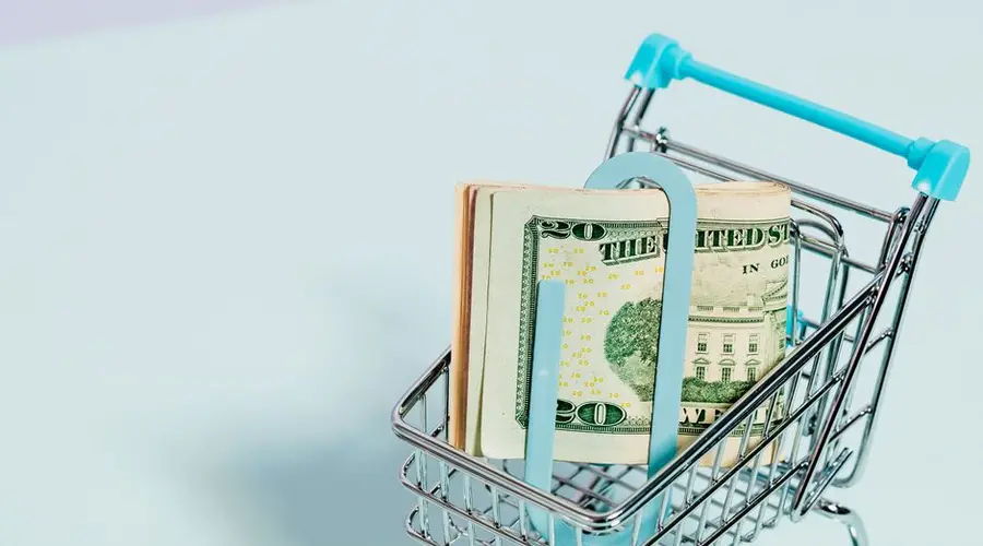 A shopping cart holding a stack of US dollar bills symbolizes financial savings and consumer spending.
