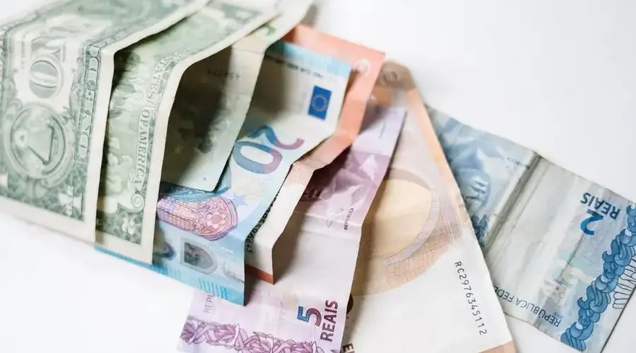 A collection of different international currency notes, including US Dollar, Euro, and Brazilian Real, on a white background.