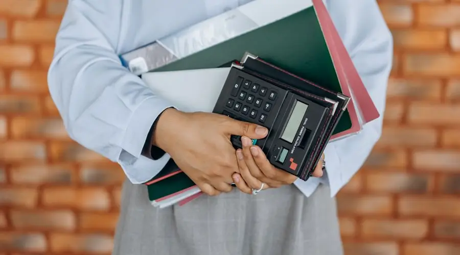 Close-up of a person holding a calculator and folders against a brick wall background.