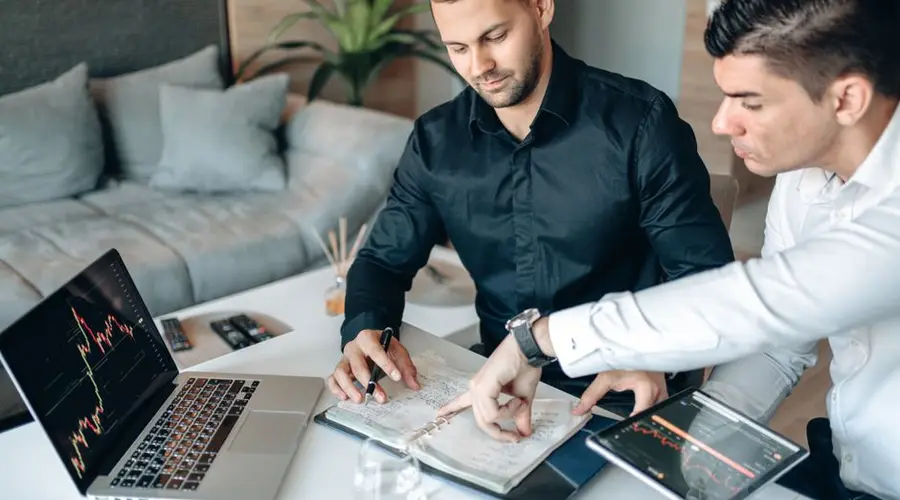 Two businessmen collaborating on stock market analysis with charts on laptops and tablets in a modern office.