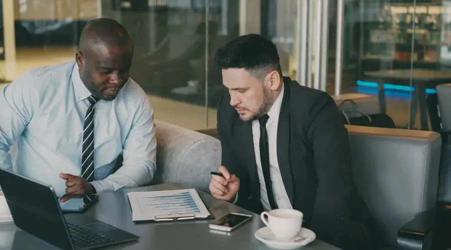 Two businessmen reviewing a financial report with laptop and coffee in office setting.