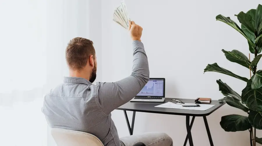 A man in a home office raises cash, symbolizing financial success and achievement.