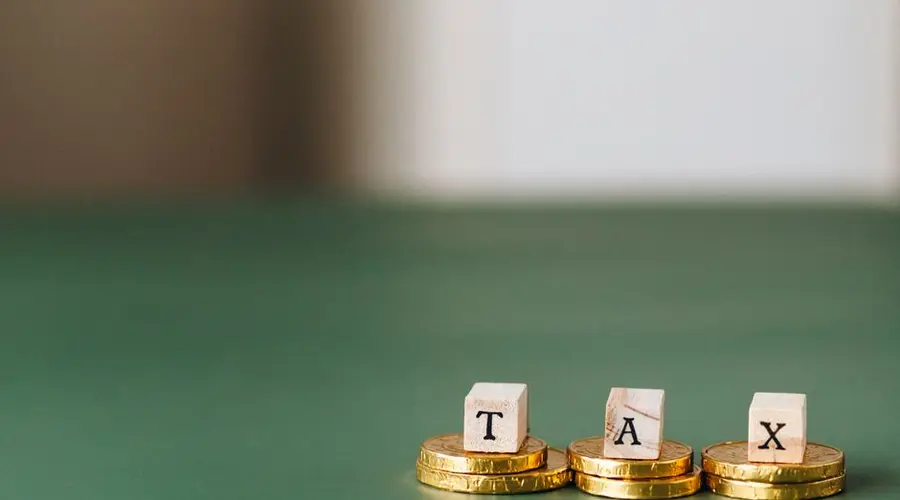 Stacked gold coins with wooden blocks spelling 'TAX' on a green backdrop.