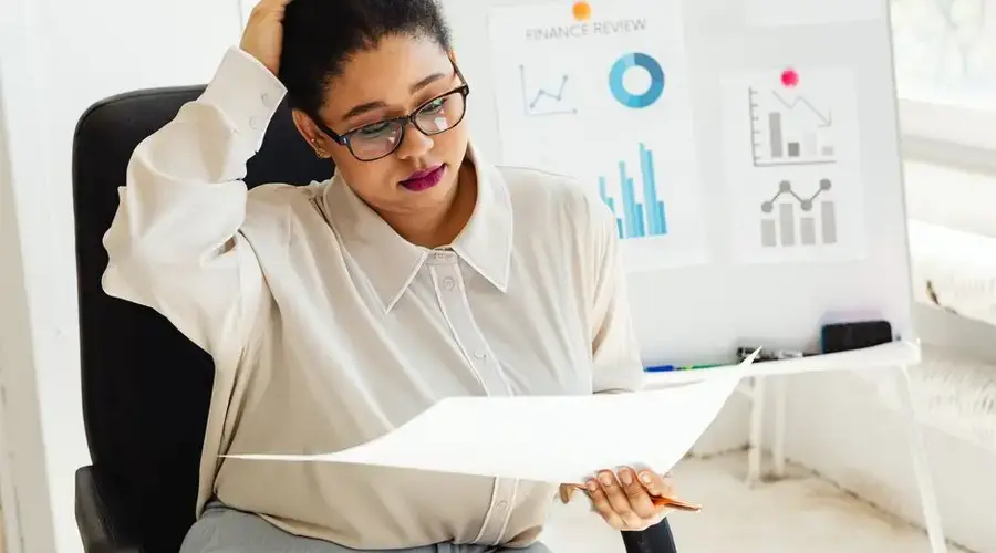 An adult woman examines financial reports at her office desk with charts and graphs in the background.