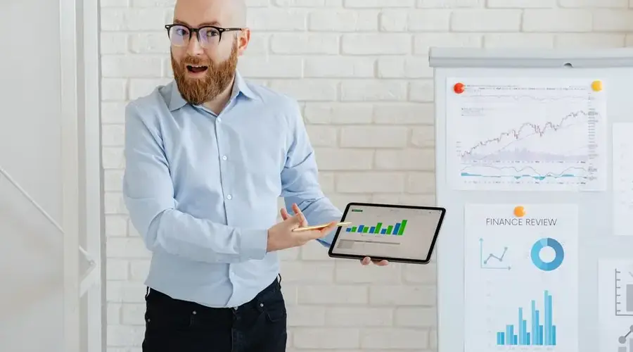 Bearded man in glasses presenting financial charts on tablet indoors.