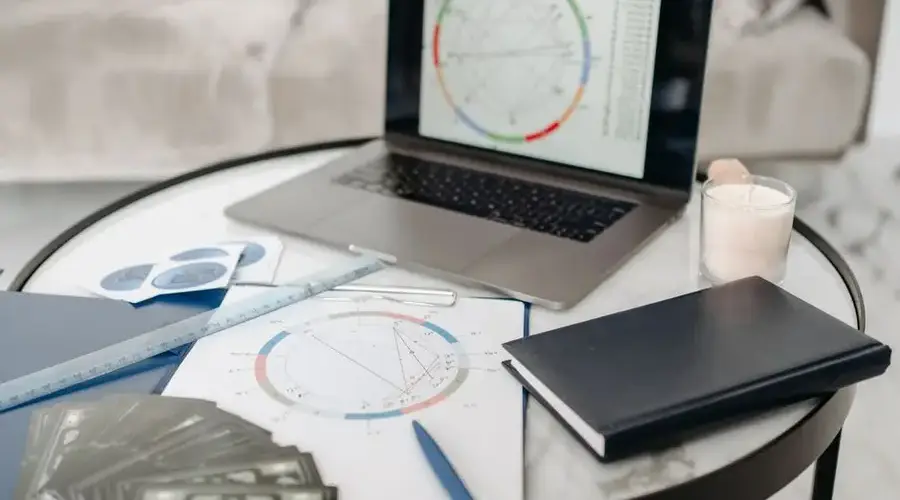 Astrology charts displayed on a laptop next to tarot cards and notebooks on a table.