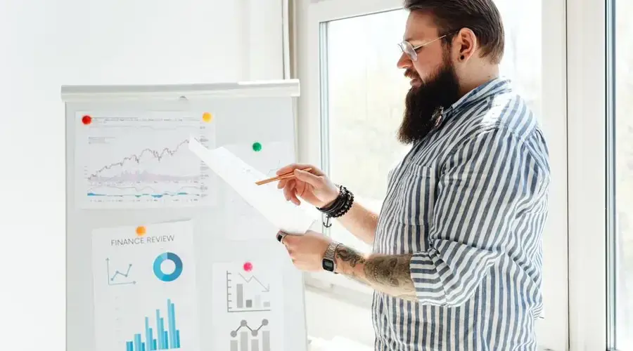 A bearded man in glasses reviews financial charts on paper next to a whiteboard.
