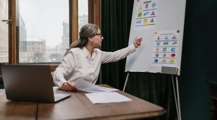 Senior woman conducting an online geometry lesson, highlighting various shapes on a chart with a laptop.