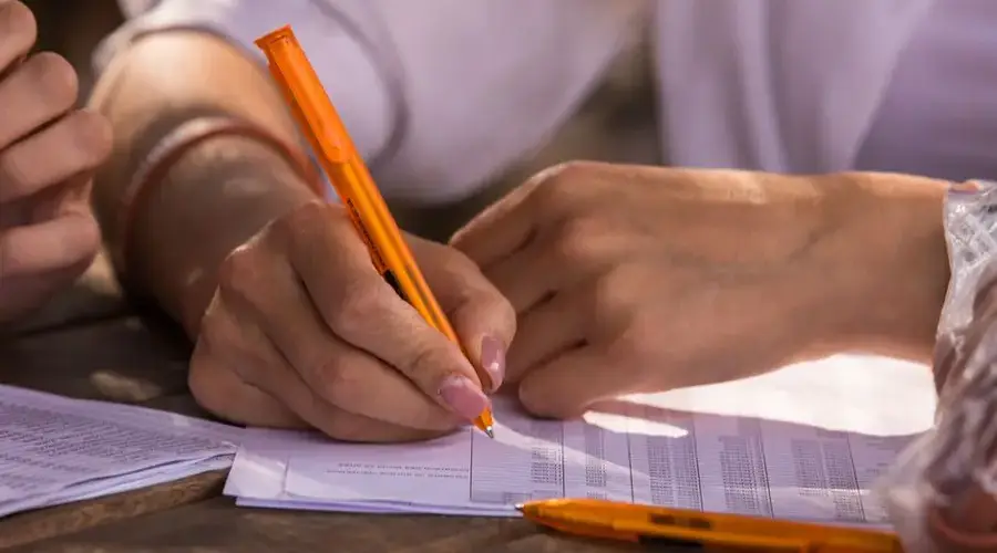 Close-up shot of hands writing on documents with orange pen, implying business or education setting.