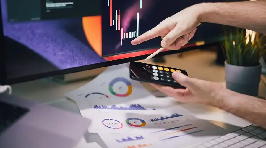 Hands analyzing stock market data on monitor and smartphone in office setting.