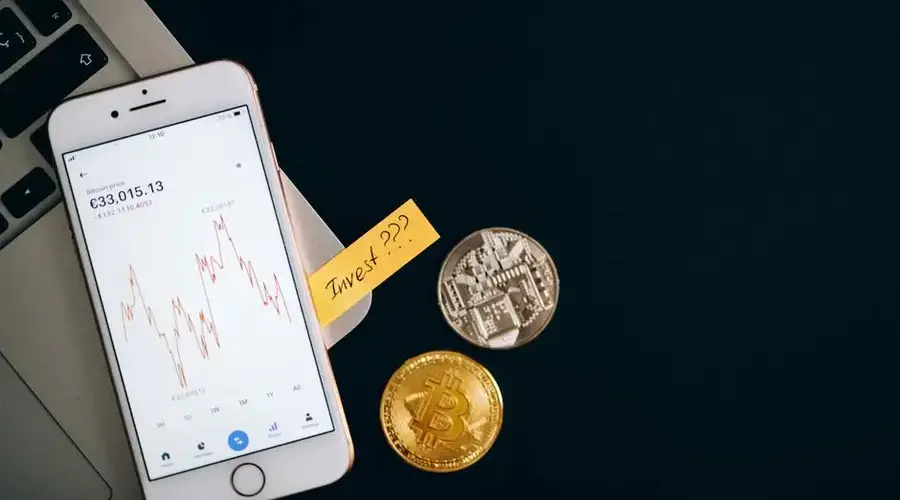 A smartphone displaying cryptocurrency prices beside coins and a laptop, symbolizing investment.