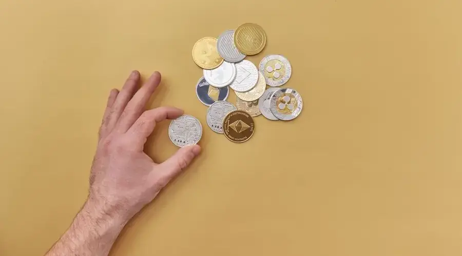 A hand picks cryptocurrencies from a mix of gold and silver coins on a vibrant yellow background.