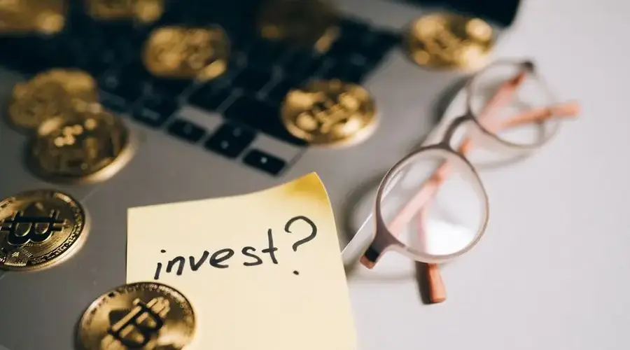 A close-up of bitcoin coins on a laptop with eyeglasses and a sticky note reading 'invest?'