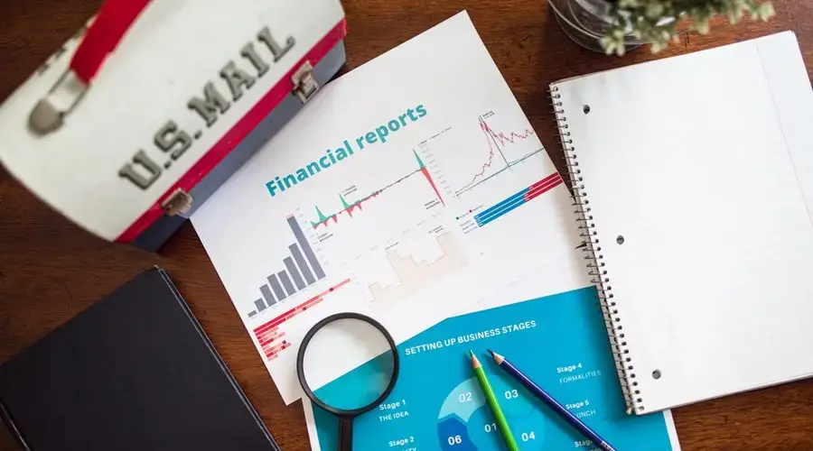 Financial charts and office supplies on a wooden desk, featuring a magnifying glass and US Mail lunchbox.