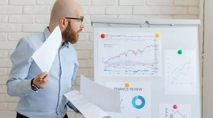 Bald businessman in smart casual attire analyzing financial charts on a whiteboard in an office setting.