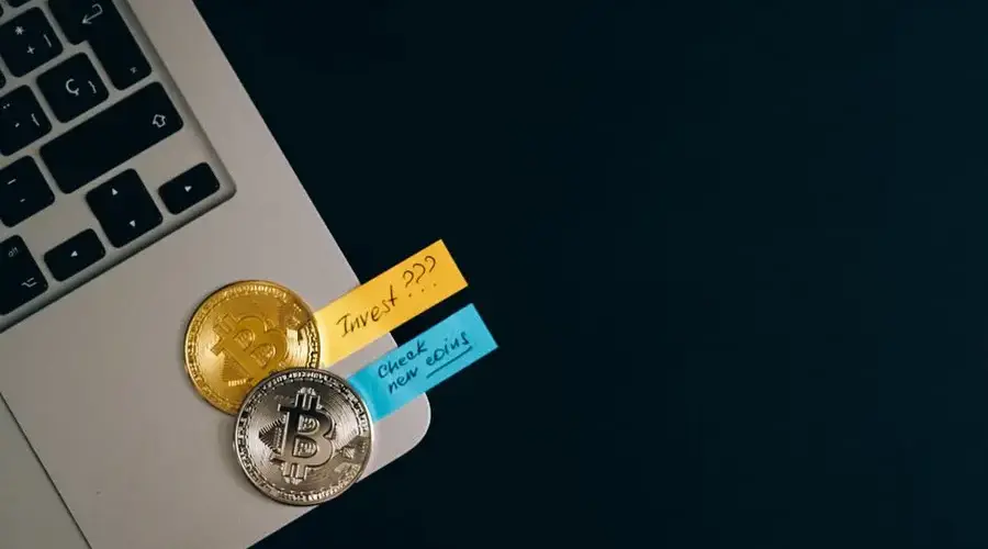 Bitcoin coins placed on a laptop keyboard with sticky note reminders about investing and checking new coins.
