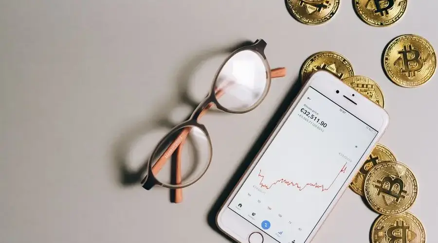 Flat lay of Bitcoin coins, eyeglasses, and smartphone displaying cryptocurrency graph.