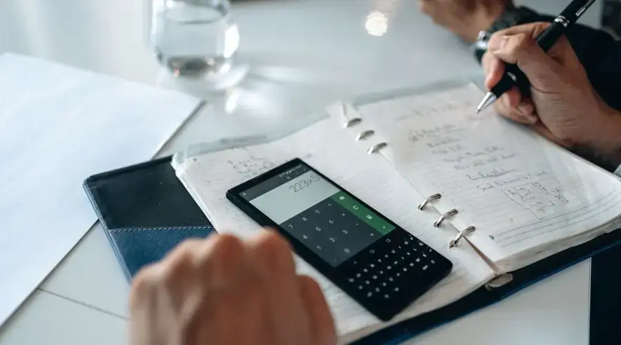 Close-up of hands writing in notebook with smartphone calculator on the table.