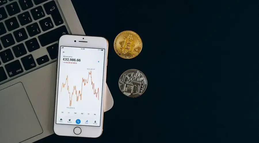 Smartphone displaying Bitcoin price chart alongside Bitcoin and Ethereum coins on black background.