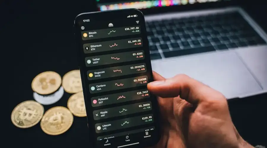 A person checks cryptocurrency values on a smartphone with a laptop and Bitcoins nearby.
