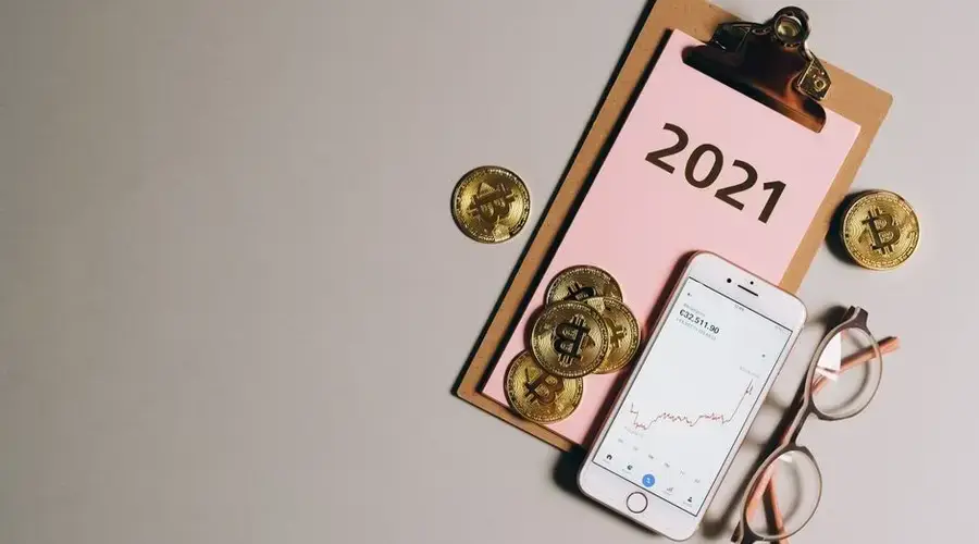 Flat lay of bitcoins, smartphone, eyeglasses, and clipboard with 2021 text, symbolizing cryptocurrency trends.